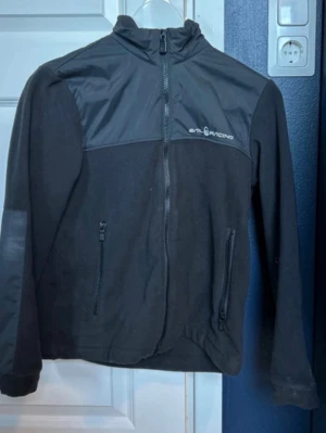 Svart vindjacka från Evil Racing - Snygg svart vindjacka från Evil Racing med hög krage och dragkedja framtill. Jackan har två fickor med dragkedja och detaljer i blankare material över axlar och bröst. Perfekt för dig som gillar sportig stil och vill ha en lätt jacka till vardagen. Storlek 152-156