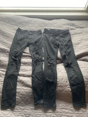 Svarta slim jeans med slitningar  - Två par svarta slim jeans med slitningar och hål på framsidan av benen. Jeansen har klassisk femficksmodell och låg midja. Perfekta för en edgy och avslappnad stil. Materialet är troligtvis bomull med stretch för bekväm passform.