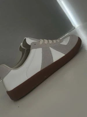 Maison Margiela Gats  - Snygga sneakers i vitt och ljusgrått med klassisk design. Ovandelen är i slätt läder och mocka, och skorna har en mörkbrun gummisula. Ljusbeige snörning och diskreta detaljer ger en clean look som passar till det mesta. De är endast använda en gång och priset är ej hugget i sten och kan diskuteras.