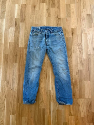 Levi's ljusblå raka jeans - Säljer ett par klassiska ljusblå Levi's jeans med rak passform och fem fickor. Jeansen har en vintage look med slitningar och den ikoniska läderlappen bak i midjan. Perfekta för en avslappnad och trendig stil.