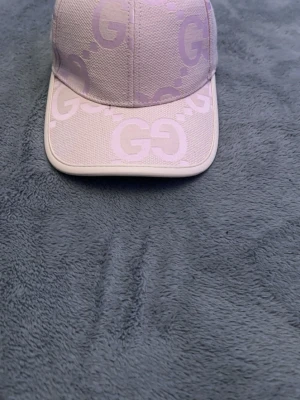 Ljusrosa keps från Gucci - Säljer en snygg ljusrosa keps från Gucci med det klassiska GG-mönstret över hela kepsen. Kepsen har böjd skärm och justerbart band bak. Tillverkad i bomullsmix och har en exklusiv känsla. Perfekt för dig som vill ha en trendig accessoar med lyxig touch.