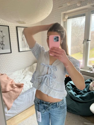 Ljusblå blus med knyt och volang - Supersöt ljusblå blus med två knyt framtill, v-ringning och volangkant. Puffiga korta ärmar med resår och lätt croppad passform. Perfekt för dig som gillar romantiska detaljer och en feminin stil. Liten i storleken.