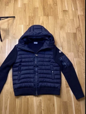 Svart pufferjacka med huva från Moncler - Snygg svart pufferjacka från Moncler med stickade ärmar och vadderad kropp. Jackan har en stor huva, två fickor med tryckknappar och dragkedja framtill. Moncler-logga på vänster ärm och ribbade muddar vid ärmslut och nederkant. Perfekt för kalla dagar.