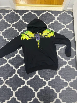 Svart hoodie Marcelo Burlon med tryck - Säljer en svart hoodie från Marcelo Burlon med färgstarkt tryck i gult och grönt över axlarna och bröstet. Hoodien har huva med snörning och en klassisk känguruficka framtill. Perfekt för dig som vill sticka ut med en unik design.