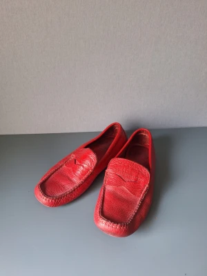 Röda loafers i skinn från Hush puppies - Sköna loafers från Hush puppies.Säljer ett par klassiska röda loafers i mjukt skinn med stilren design och markerade sömmar. Skorna har en tidlös pennyloafers-look och är perfekta för dig som vill sticka ut med färg. Passar till många olika outfits och ger en cool touch till din stil. Observera att skinnet är nött på sulan under stortån, vilket syns tydligt på bild. Här är skinner tjockt så det kommer inte vara något problem för funktionen.
