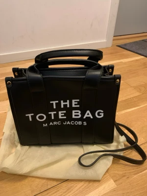 Svart The Tote Bag  - Snygg svart handväska med texten 'THE TOTE BAG' Marc Jacobs  i vitt på framsidan. Väskan har både handtag och en lång axelrem, samt guldfärgade detaljer. Tillverkad i slätt konst läder med en boxig form och diskret logga på baksidan.inköpt utomlands Inget kvitto finns el äkthetsbevis  så inga garantier gällande märket säljes därför billigt. Väskan är helt oanvänd. Vid frågor skriv gärna.