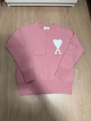Rosa stickad tröja från Ami - Säljer en rosa stickad tröja från Ami med rund halsringning och ribbade muddar. På bröstet finns ett vitt hjärta och bokstaven A som ger en unik touch. Perfekt för dig som gillar stilrena men ändå lekfulla plagg.