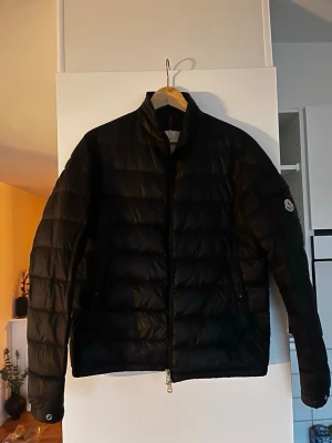 Svart pufferjacka från Moncler - Säljer en svart pufferjacka från Moncler med klassisk quiltad design och hög krage. Jackan har dragkedja framtill, två sidofickor och Moncler-logga på ärmen. Perfekt för kalla dagar och riktigt snygg till streetwear-stilen.