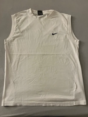 Vit ärmlöst linne från Nike - Säljer ett clean vitt linne från Nike med klassisk svart logga på bröstet. Linnet är ärmlöst och har rund halsringning. Perfekt för dig som gillar sportig och enkel stil. Materialet är mjuk bomull som känns skönt mot huden.