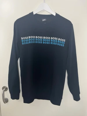 Svart sweatshirt från BOSS - Snygg svart sweatshirt från BOSS med tryckt logga i vitt och blått över bröstet. Tröjan har rund halsringning och långa ärmar. Perfekt för en avslappnad och trendig stil.
