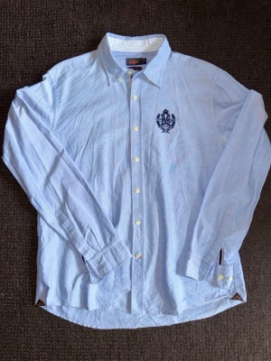 Ljusblå skjorta från Morris - Fin button-down skjorta från Morris med broderat emblem på bröstet. Storlek XL men passar bra som L. Skjortan har klassisk krage, långa ärmar och flera typiska Morris-detaljer. Tillverkad i bomull och har en clean, stilren look som passar perfekt till jeans eller chinos. Från rök- och djurfritt hem. 