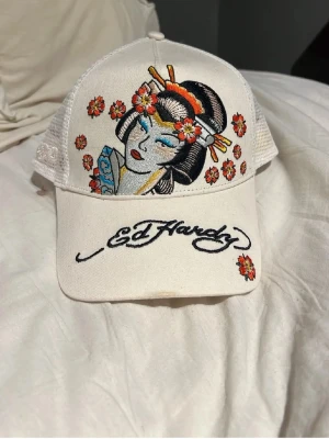 Vit Ed Hardy keps med geisha-brodyr - Snygg vit keps från Ed Hardy med broderad geisha och blommor framtill. Kepsen har nät på sidorna och bak för extra ventilation. Broderad Ed Hardy-logga på skärmen och coola färgdetaljer i rött, gult och blått. Perfekt statement-piece för din outfit.