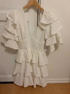 NY! Vit volangklänning med v-ringning - Supersöt vit klänning med flera lager volanger och djup v-ringning. Endast testad och har prislappen kvar men gått för lång tid för att retunera. Klänningen har kort längd och vida, volangprydda ärmar som ger en romantisk känsla. Perfekt för studenten eller sommarens alla tillfällen! Jag är strax under 170 och skulle säga att den är något längre än ”normalt”. Extra bra längd för studenten!