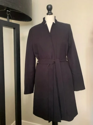 Svart klassisk kappa med knytband - Stilren svart kappa med rak passform och knytband i midjan. Kappan har ståkrage, dolda knappar framtill och långa ärmar. Perfekt för dig som gillar minimalistisk och tidlös stil. Passar till många olika outfits och är enkel att matcha.