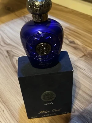 Lattafa Blue Oud Eau de Parfum - Lattafa Blue Oud Eau de Parfum kommer i en lyxig, djupblå flaska med guldfärgade detaljer och ett elegant mönstrat lock. Förpackningen är svart med silverfärgade inslag och stilren design. Flaskan rymmer 100 ml och har en exklusiv känsla.