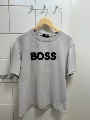 Grå BOSS t-shirt med tryck - Snygg grå t-shirt från BOSS med stort svart BOSS-tryck på bröstet. Klassisk rund halsringning och korta ärmar. Tillverkad i mjuk bomull för en bekväm känsla och enkel stil.