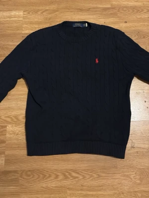 Mörkblå kabelstickad tröja Polo Ralph Lauren - Kabelstickad mörkblå tröja från Polo Ralph Lauren med rund halsringning och röd broderad logga på bröstet. Tröjan har ribbade muddar vid ärmslut och nederkant. Perfekt för dig som gillar klassisk och stilren design.