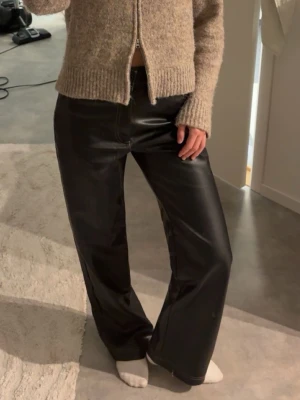 Svarta vida jeans i skinnimitation - Säljer ett par svarta jeans i skinnimitation med raka, vida ben och hög midja. Jeansen har klassiska fickor fram och bak samt kontrastsömmar. Perfekta för dig som vill ha en edgy och trendig look.