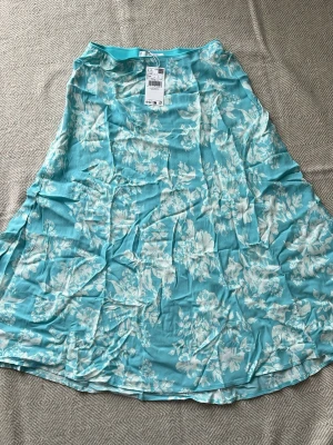 Blommig turkos långkjol Mango MNG - Snygg turkos långkjol med vitt blommönster. Kjolen har en luftig och flowy passform, perfekt för varma dagar. Tillverkad i ett lätt och mjukt material som ger en skön känsla. Passar dig som vill ha en fräsch och somrig look.