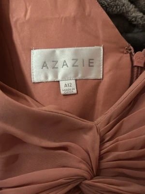 Lång baby rosa klänning från Azazie - Säljer en elegant långklänning från Azazie i en baby rosa ton. Klänningen har v-ringad hals, breda axelband och ett twistat knyt framtill. Tillverkad i ett mjukt, flytande material som ger ett snyggt fall. Perfekt för speciella tillfällen.