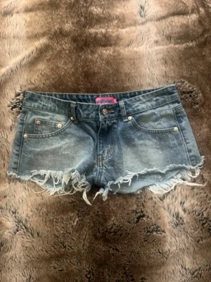 Blå jeansshorts med fransar - Säljer ett par blå denimshorts med slitna, fransiga kanter och klassisk femficksdesign. Shortsen har låg midja och bälteshällor, perfekta för en avslappnad och trendig sommarlook.