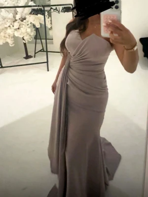 Ljuslila långklänning med drapering - Elegant ljuslila långklänning med figurnära passform och vacker drapering över midjan. Klänningen har axelbandslös design och ett långt släp som ger en lyxig känsla. Perfekt för speciella tillfällen där du vill sticka ut. Balklänning festklänning 