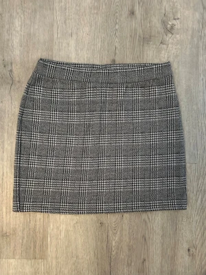 Grå rutig kortkjol - Snygg grå kortkjol med klassiskt rutigt mönster. Kjolen har en rak passform och är perfekt för dig som gillar en stilren och trendig look. Passar till både stickad tröja och t-shirt för en cool vardagsstil.