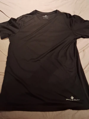 Svart tränings-tshirt från Ronhill - Säljer en svart tränings-tshirt från Ronhill. T-shirten har rund hals, korta ärmar och är tillverkad i ett lätt och snabbtorkande funktionsmaterial. Perfekt för löpning eller gym. Diskret logga i vitt nere vid fållen.