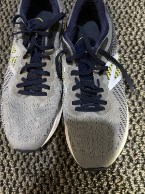 Gråa New Balance sneakers med gula detaljer - Snygga sneakers från New Balance i grå mesh med mörkblå snörning och innersula. Skorna har vita och gula detaljer på sidan och hälen samt en vit sula. Perfekta för dig som gillar sportig stil och vill ha lätta skor med bra ventilation.