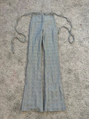 Grå rutiga leggings med knytband - Snygga grå leggings med rutigt mönster och långa knytband i midjan. De har en smal passform och är utsvängda nedtill, vilket ger en cool retrostil. Perfekta för dig som vill ha något trendigt och bekvämt. Materialet är mjukt och stretchigt.