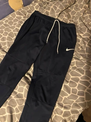 Svarta Nike mjukisbyxor med snörning - Snygga svarta mjukisbyxor från Nike med vit dragsko i midjan och klassisk vit Nike-logga på låret. Byxorna har en avslappnad passform och är perfekta för chill dagar. Tillverkade i mjukt material som känns skönt mot huden.