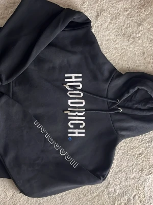 Svart hoodie från Hoodrich - Snygg sprillans ny svart hoodie från Hoodrich med vit logga tryckt vertikalt på bröstet och detaljer på fickan. Klassisk känguruficka och justerbar huva med snören. Perfekt för en streetwear-look och riktigt bekväm att bära. Endast provad.