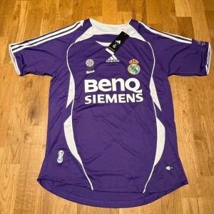 Real Madrid third kit 06/07 X R.Carlos #3 - Säljer en lila Real Madrid matchtröja från Adidas med vita detaljer och tryck. Tröjan har Benq Siemens sponsorlogga på framsidan, klubbmärke och nummer 3 med R. Carlos på ryggen. Perfekt för fotbollsfans som vill sticka ut på planen.
