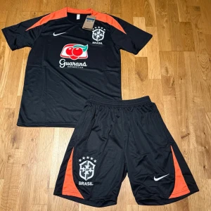 Brazil summer set svart & orange - Säljer ett svart träningsset från Nike med Brasil-logga. Setet består av en t-shirt och shorts med orange detaljer på axlar och sidor. Materialet är lätt och andas, perfekt för fotboll eller träning. Guaraná Antarctica sponsortryck på tröjan. Elastisk midja på shortsen.