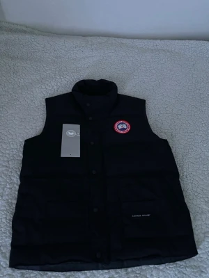 Svart dunväst från Canada Goose - Snygg svart dunväst från Canada Goose med hög krage och tryckknappar framtill. Västen har det klassiska Canada Goose-märket på bröstet och två stora fickor framtill. Perfekt för lager-på-lager och håller dig varm med sin vadderade design.