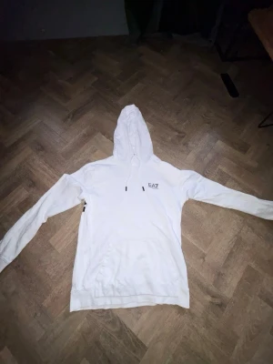 Vit hoodie från EA7 Emporio Armani - Snygg vit hoodie från EA7 Emporio Armani med svart logga på bröstet och coolt svartvitt mönster på ryggen. Klassisk känguruficka framtill och justerbar huva med snören. Size: S. Mer tröjor finns i min profil