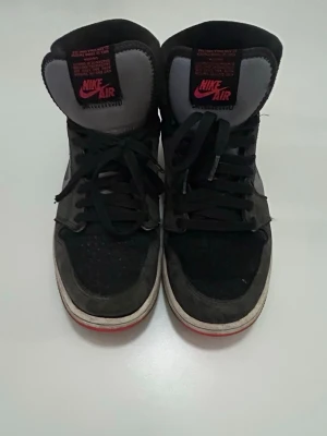 Nike Air Jordan 1 High svart/röd - Nike Air Jordan 1 High sneakers i svart mocka och läder med grå detaljer och röd sula. Klassisk high-top siluett med svart snörning och ikonisk Nike Air-logga på plösen. Perfekta för dig som gillar streetwear och vill sticka ut.