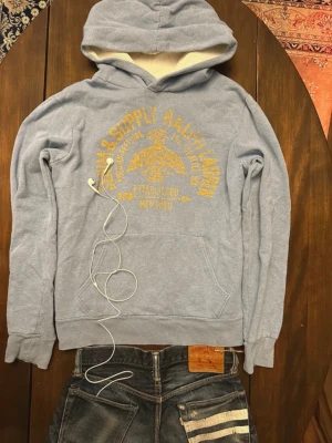Ralph Lauren Denim&Supply - Ljusblå hoodie från Denim&Supply Raplhlauren med gul vintageinspirerad text och tryck på bröstet. Känguruficka och klassisk huva. Perfekt för en avslappnad och cool stil. Materialet känns som bomullsblandning.