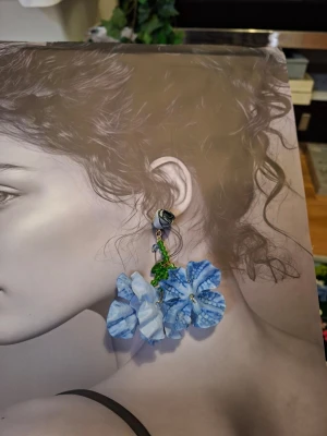 Blå blomformade statementörhängen - Unika örhängen med stora blå blommor i tyg och gröna pärldetaljer. Fästet är utformat som en liten grå ros. Perfekta för dig som vill sticka ut med färg och form. Materialet är mestadels textil och plast.