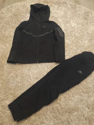 Svart Nike träningsset med huva - Säljer ett svart träningsset från Nike med zip hoodie och matchande byxor. Hoodien har dragkedja, huva och diskreta reflexdetaljer på bröstet och ärmen. Byxorna har resår i midjan och smal passform. Båda plaggen har Nike-logga och är tillverkade i mjukt, stretchigt material.