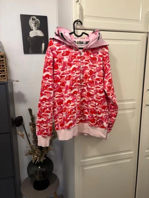 BAPE rosa camo hoodie med hajmotiv - Säljer en BAPE hoodie med ikoniskt rosa och rött camo-mönster samt hajmotiv på huvan. Tröjan har dragkedja, ribbade muddar och fickor framtill. Perfekt för dig som gillar streetwear och vill sticka ut med en unik design.