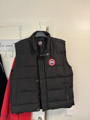 Svart dunväst från Canada Goose - Snygg svart dunväst från Canada Goose med klassisk logga på bröstet. Västen har hög krage, två framfickor med dragkedja och tryckknappar framtill. Perfekt för lager på lager och håller dig varm under kyliga dagar.