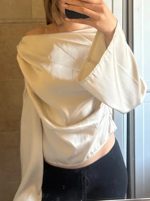 Offshoulder vit omlottopp - Säljer en vit offshoulder omlottopp med vida ärmar. Toppen har en lös passform och är tillverkad i ett mjukt, glansigt material som ger en lyxig känsla. Perfekt för dig som vill ha en unik och trendig look.