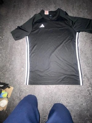 Svart Adidas sporttröja med vita detaljer - Säljer en svart sporttröja från Adidas med vita ränder längs sidorna och den klassiska Adidas-loggan på bröstet. Tröjan har rund hals och är tillverkad i ett lätt och snabbtorkande material, perfekt för träning eller match.