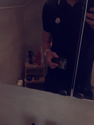 Svart Moncler piké med rödvit detalj - Snygg svart pikétröja från Moncler med klassisk krage och knappar framtill. Tröjan har korta ärmar och detaljer i rött och vitt på krage och ärmslut samt Moncler-logga på bröstet. Perfekt för en clean och stilren look.