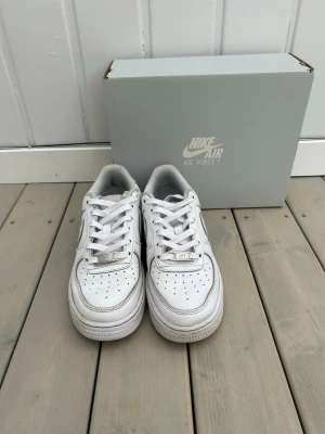 Nike Air Force 1 vita sneakers - Klassiska vita Nike Air Force 1 sneakers med låg profil och ikonisk design. Skorna har perforerad tå, vit läderöverdel, vita skosnören och en robust sula med Air-dämpning. Perfekta för dig som gillar stilrena och tidlösa sneakers.