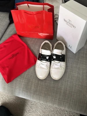 Valentino Garavani vita sneakers - Snygga vita sneakers från Valentino Garavani med svart band över vristen och nitar på sidan. Skorna har en klassisk låg silhuett och är tillverkade i läder. Perfekta för dig som vill ha en stilren och exklusiv look. Säljer skorna pga att de har blivit små.