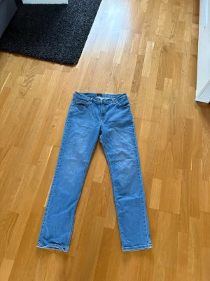 Blå raka jeans med klassisk passform - Säljer ett par ljusblå jeans med rak passform och femficksdesign. Jeansen har normal midja och klassiska detaljer som metallknapp och dragkedja. Materialet är denim i bomull och de har en enkel, tidlös look som funkar till många stilar.