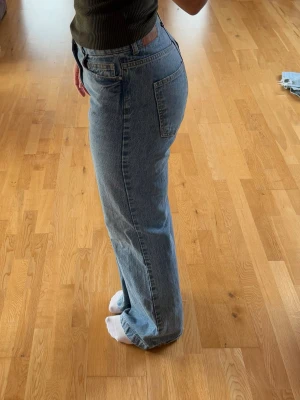 Blå raka jeans med hög midja - Säljer ett par klassiska blå jeans med rak passform och hög midja. Jeansen har fem fickor, bälteshällor och stängs med knapp och dragkedja. Perfekta till en avslappnad stil och passar till det mesta i garderoben.