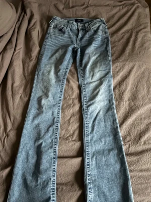 True Religion ljusblå bootcut jeans - Säljer ett par ljusblå bootcut jeans från True Religion med ikoniska rosa broderade detaljer på bakfickorna. Jeansen har klassisk femficksdesign, medelhög midja och är tillverkade i denim med snygg tvätt. Perfekta för dig som vill ha en avslappnad men trendig look. Endast använda 2 gånger, säljer då de inte kommer till användning. Inköpta för 1599.  Hör av dig ifall du har frågor🤩